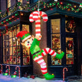 Inflatable elf in a green outfit and striped stockings climbing a red and white candy cane beside a lit house