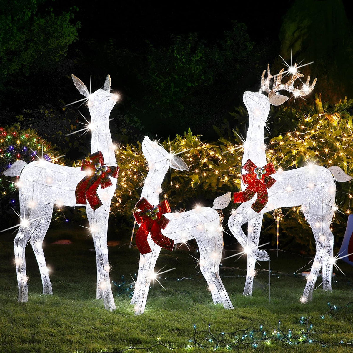 Three white lighted reindeer with red bows glowing on a lawn at night